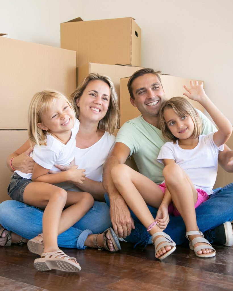 Smiling family with kids sitting on floor near cardboard boxes and relaxing. Blonde girl on father legs waving. New home owners enjoying apartment. Mortgage, relocation and moving day concept